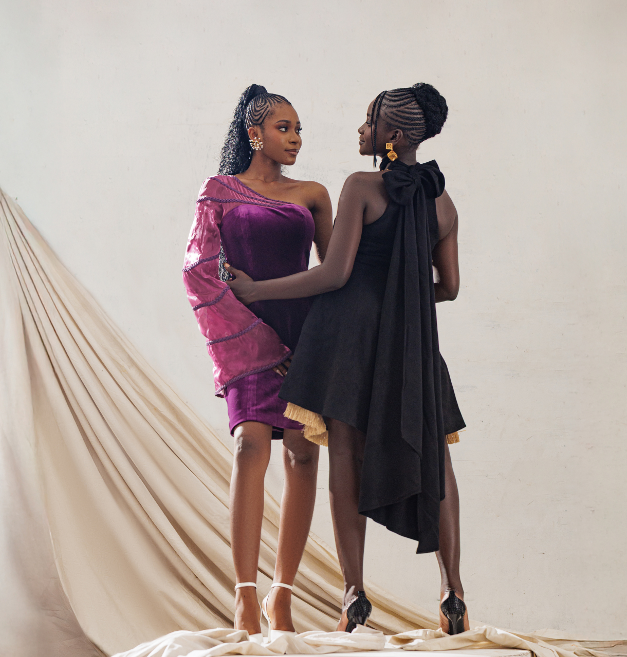 Two women in elegant dresses standing on a white platform with beige draped fabric in the background.